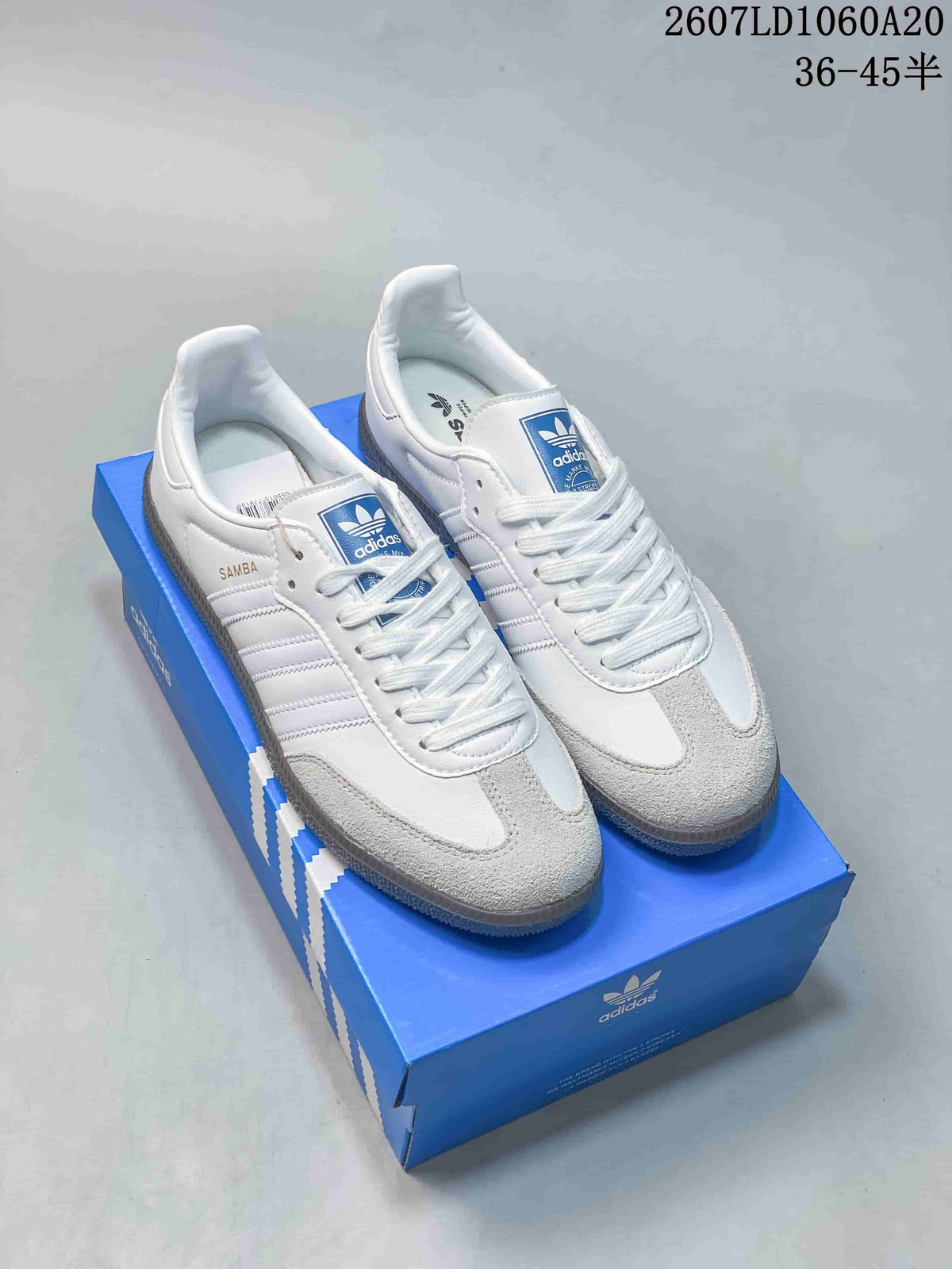 Pair of white and gray Adidas sneakers displayed on a blue shoebox, with SAMBA branding visible on the side.