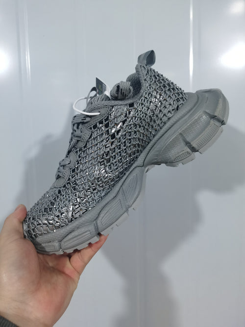 Close-up angled view of a grey and crystal fishnet-style sneaker with reflective decorative details and a segmented outsole.