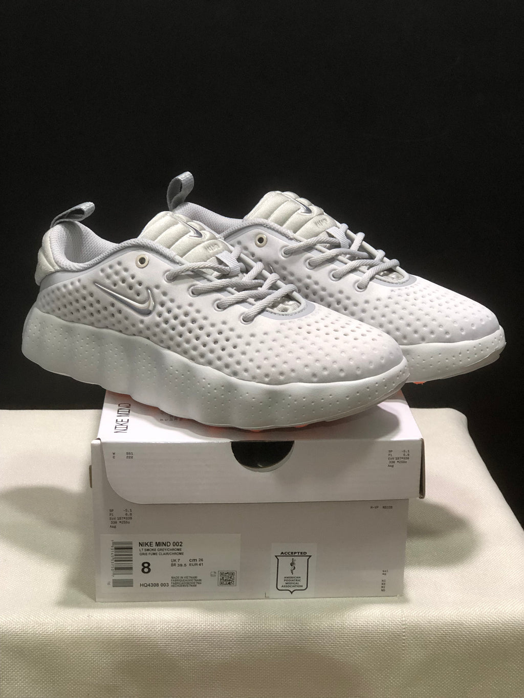 Pair of white Nike Mind 002 sneakers displayed on a product box, showing the perforated upper and sculpted sole.