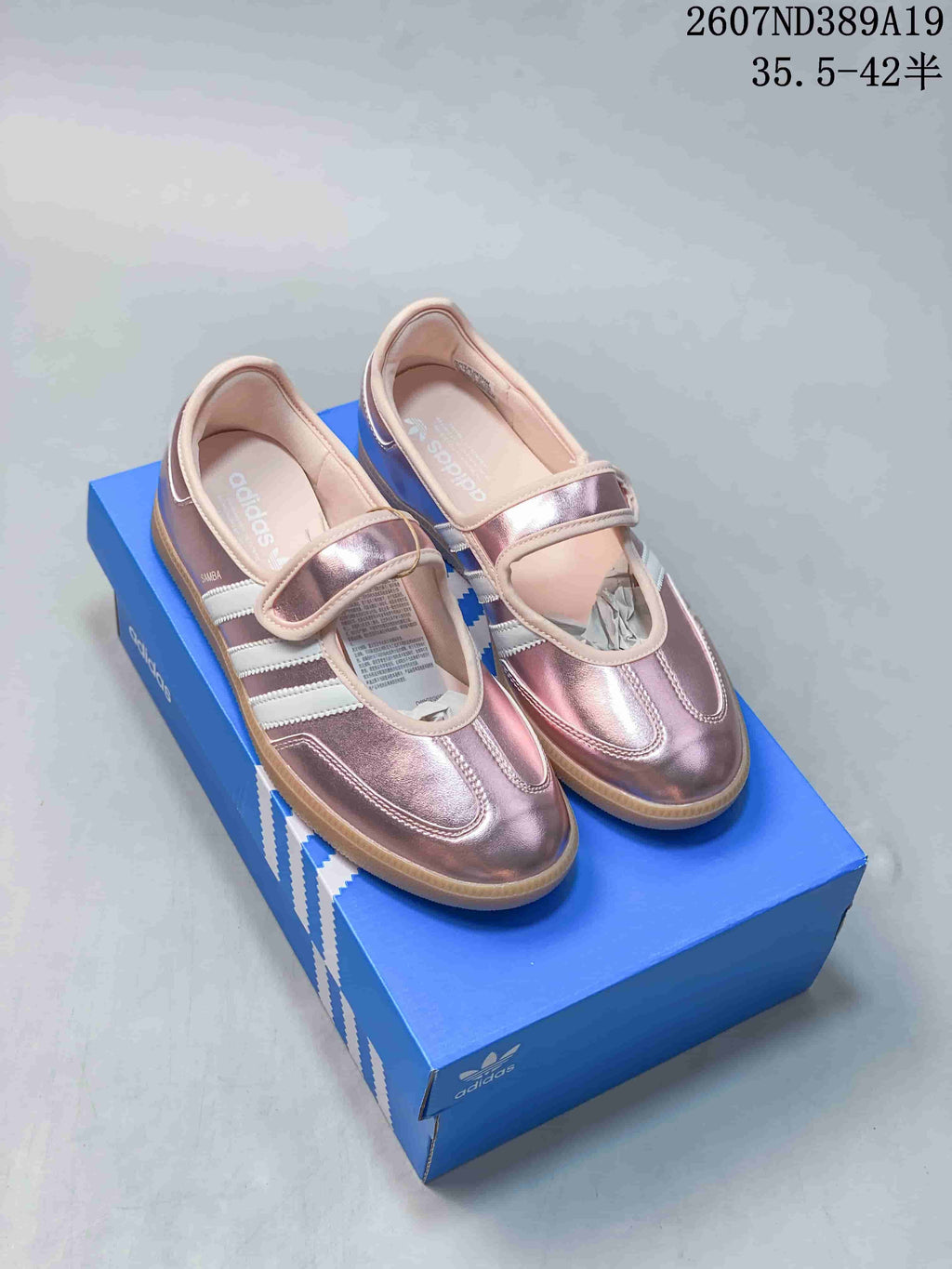 Pair of metallic pink adidas Samba Jane sneakers displayed on a blue shoebox, showing the Mary Jane straps and gum soles.