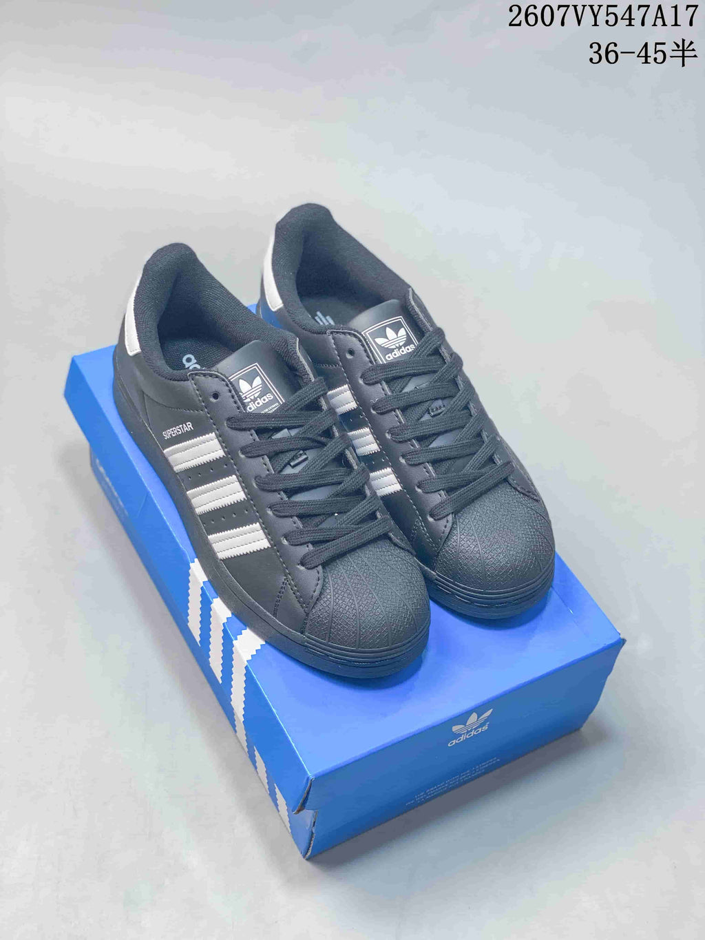 Black adidas Superstar sneakers with white stripes, black laces, and black textured toe caps displayed on a blue shoebox.