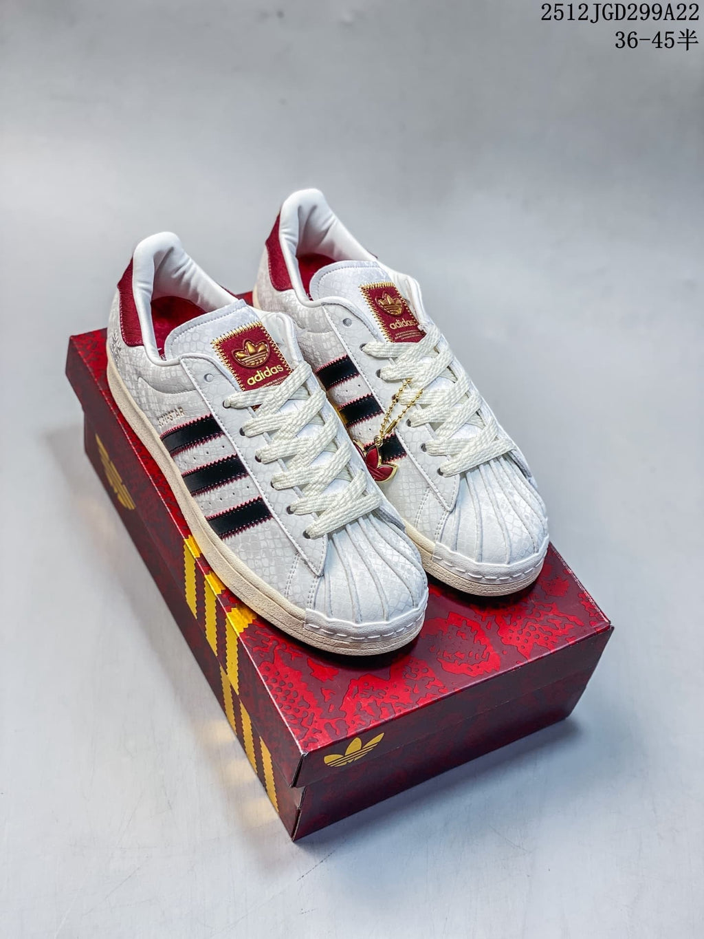 Pair of white adidas Superstar sneakers with black stripes, red accents, textured uppers, and a red patterned shoebox.