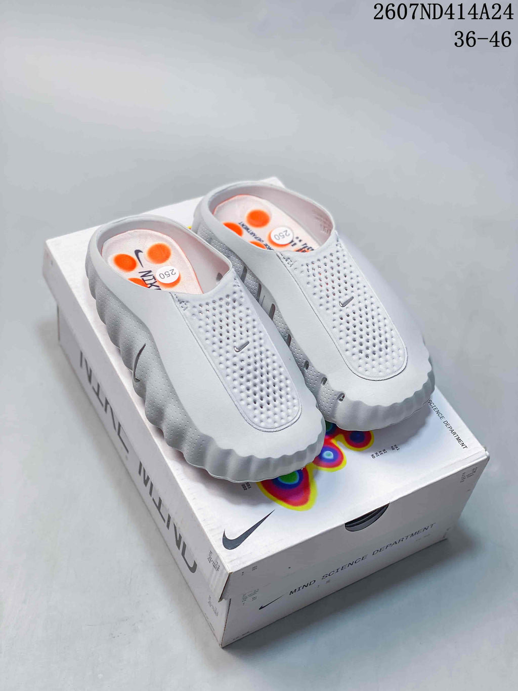 Pair of light gray Nike Mind 001 slip-on shoes displayed on a Nike Mind box, with orange insole details visible.