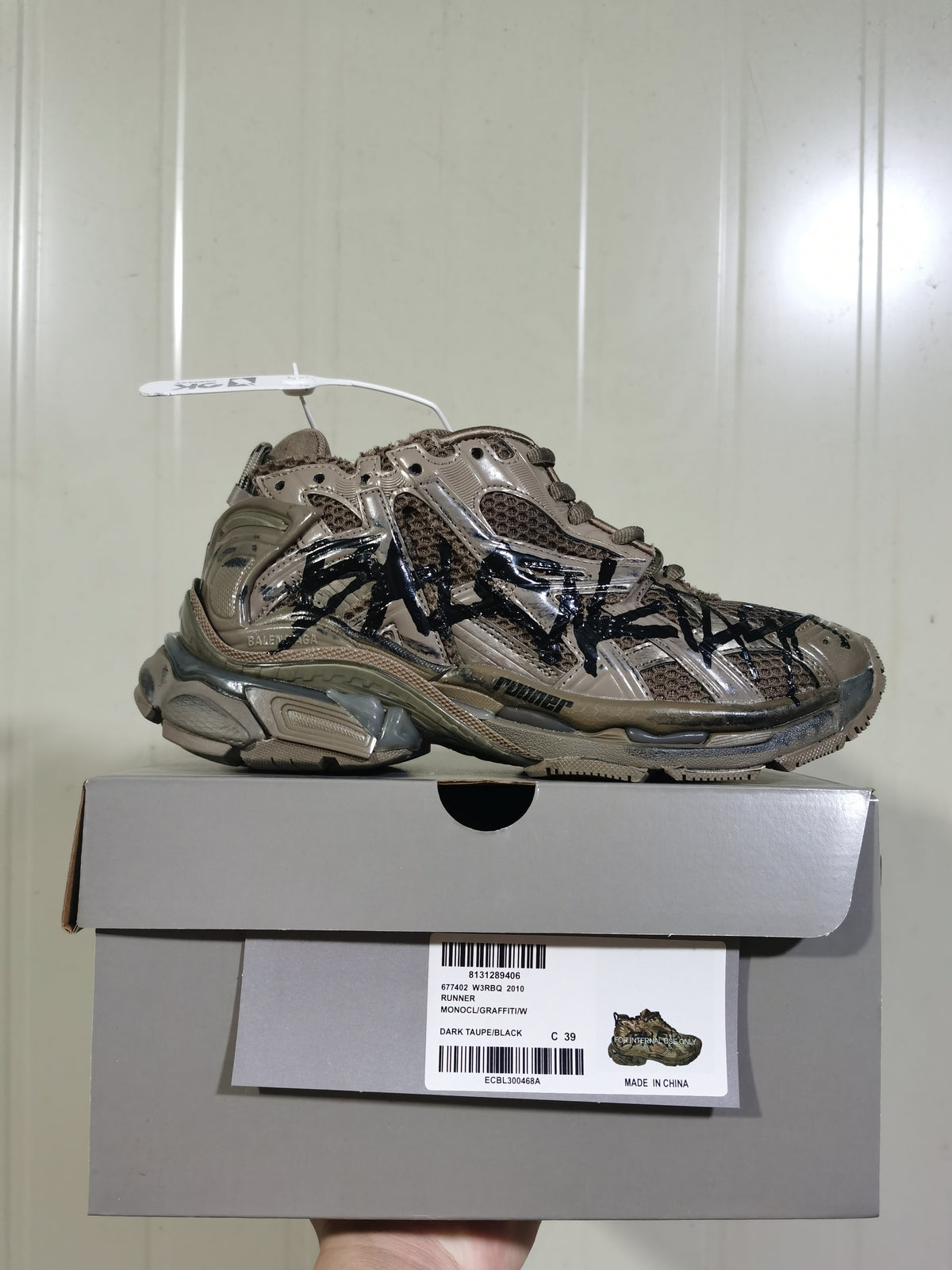 Side view of a Balenciaga Runner 2010 sneaker in dark taupe and black, displayed on its shoebox with the product label visible.