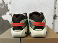 Rear view of dark mesh sneakers with green-toned overlays, cream-colored soles, and orange heel straps.