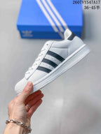 Side profile of the white low-top sneaker showing black three-stripe details, perforations, laces, and a textured shell-style toe.