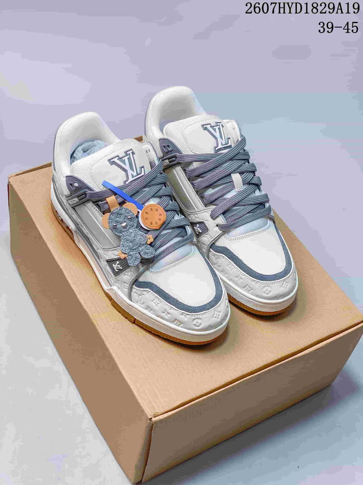 Pair of white and gray Louis Vuitton Trainer Maxi sneakers displayed on a cardboard box, with gray laces, decorative lace accessories, and the visible code 2607HYD1829A19.