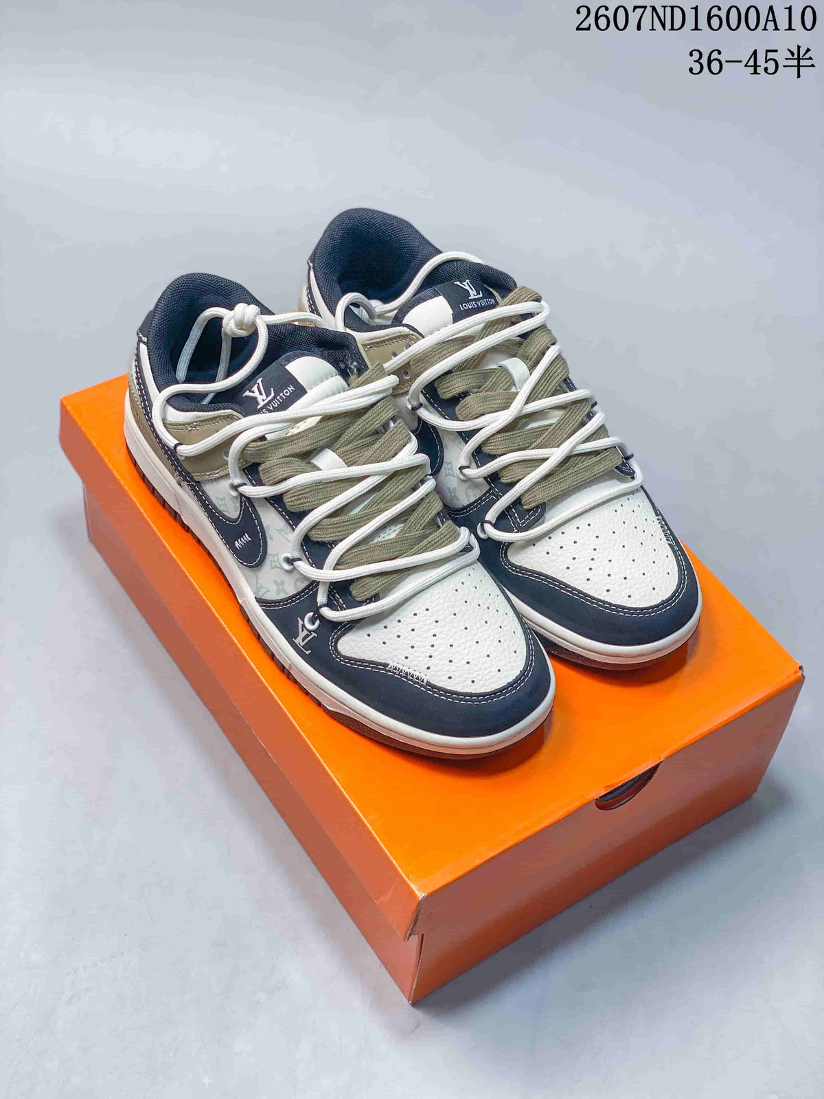Pair of low-top sneakers in navy, cream, and olive tones displayed on an orange shoebox, with multiple lace elements and a perforated toe box.