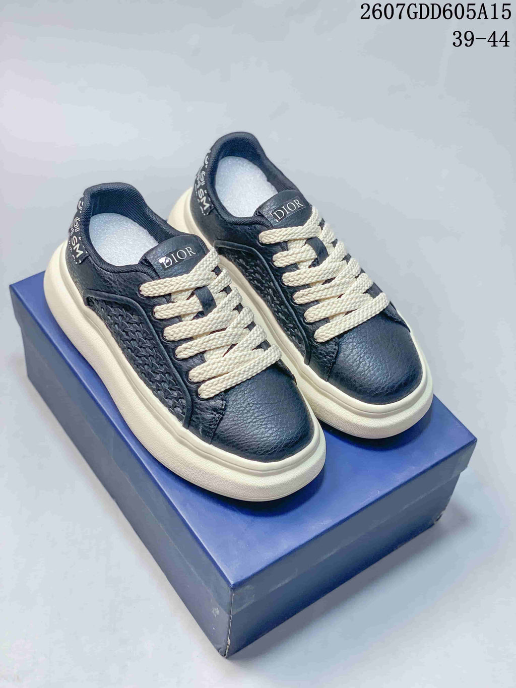 Pair of navy and white Dior low-top sneakers displayed on a blue shoebox, showing cream laces and woven side panels.