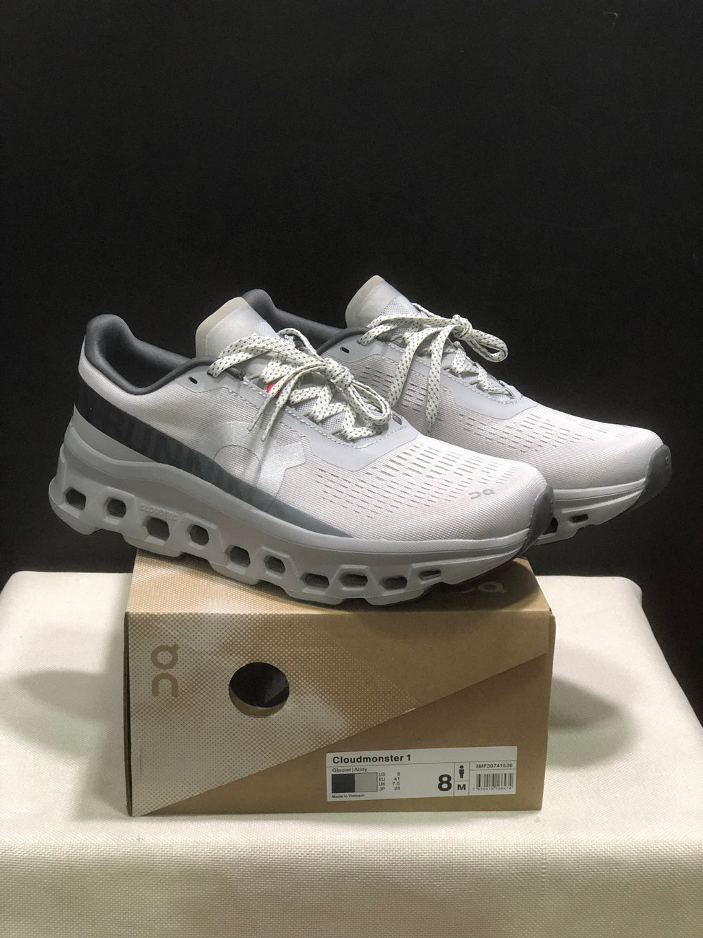 Pair of light gray and white On Cloudmonster 1 sneakers displayed on their shoebox; the box label identifies Glacier | Alloy and a photographed EU 41 sample.