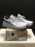 Pair of light gray and white On Cloudmonster 1 sneakers displayed on their shoebox; the box label identifies Glacier | Alloy and a photographed EU 41 sample.