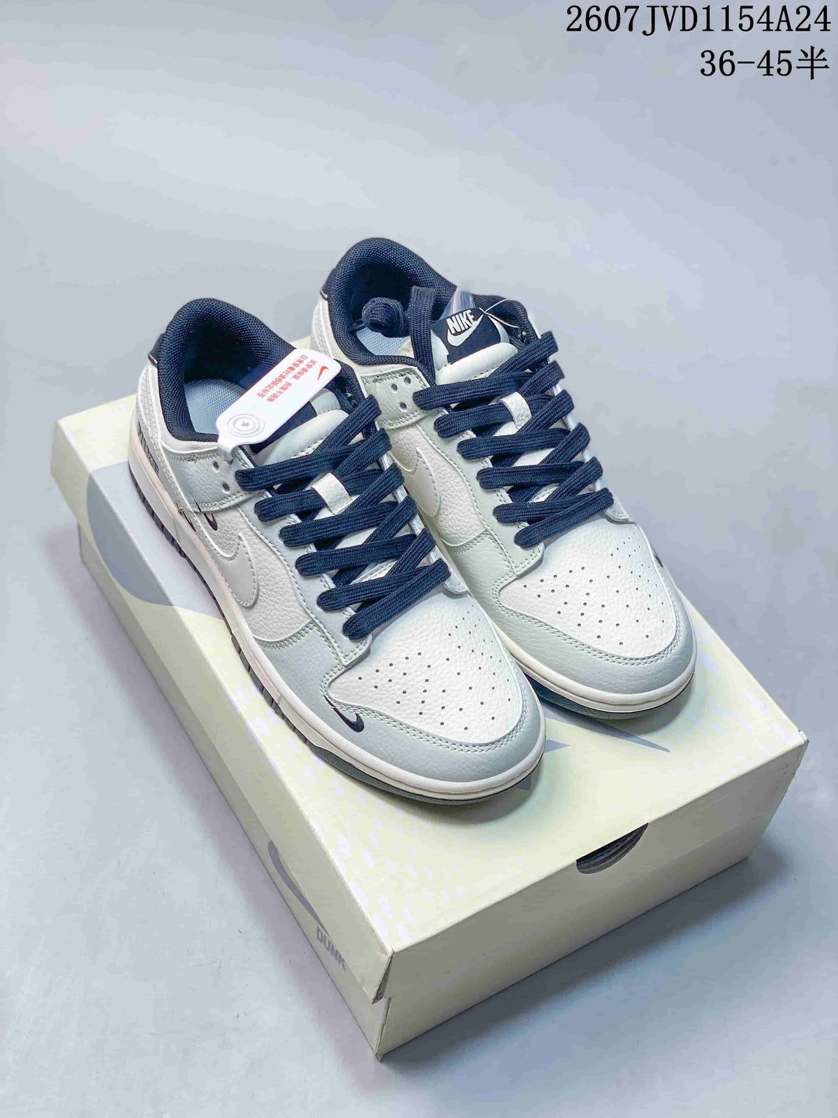 Pair of white and navy Nike SB Dunk Low sneakers displayed on a shoebox, showing navy laces, perforated toe boxes and multiple Swoosh details.