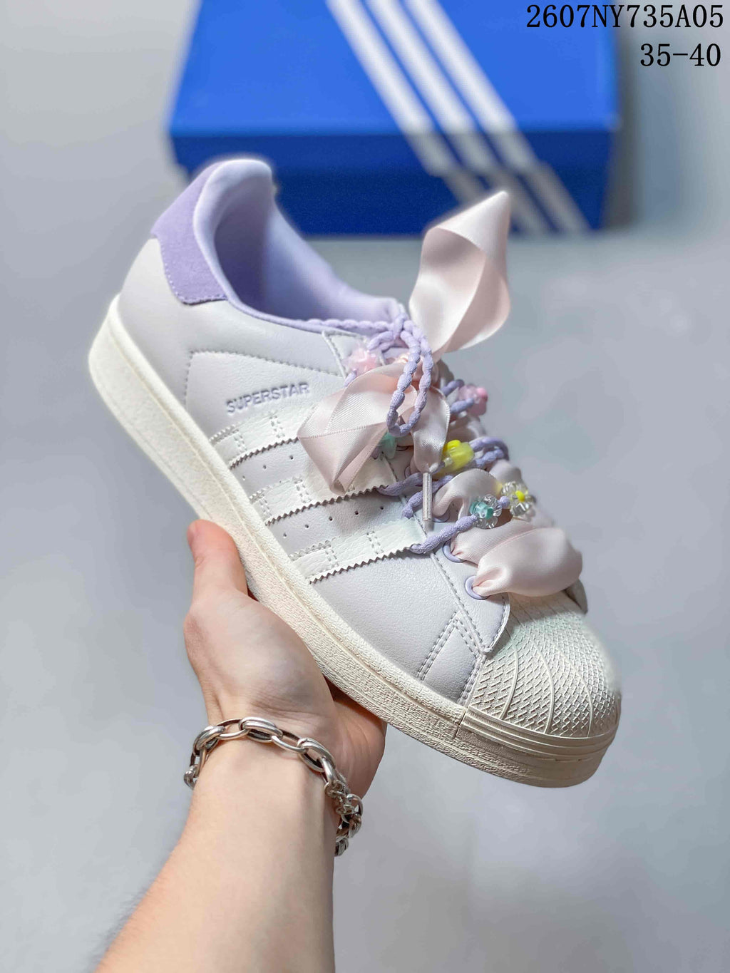 Angled side view of the white Adidas Superstar sneaker with shell toe, lilac details, and decorative ribbon laces.