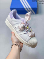 Angled side view of the white Adidas Superstar sneaker with shell toe, lilac details, and decorative ribbon laces.