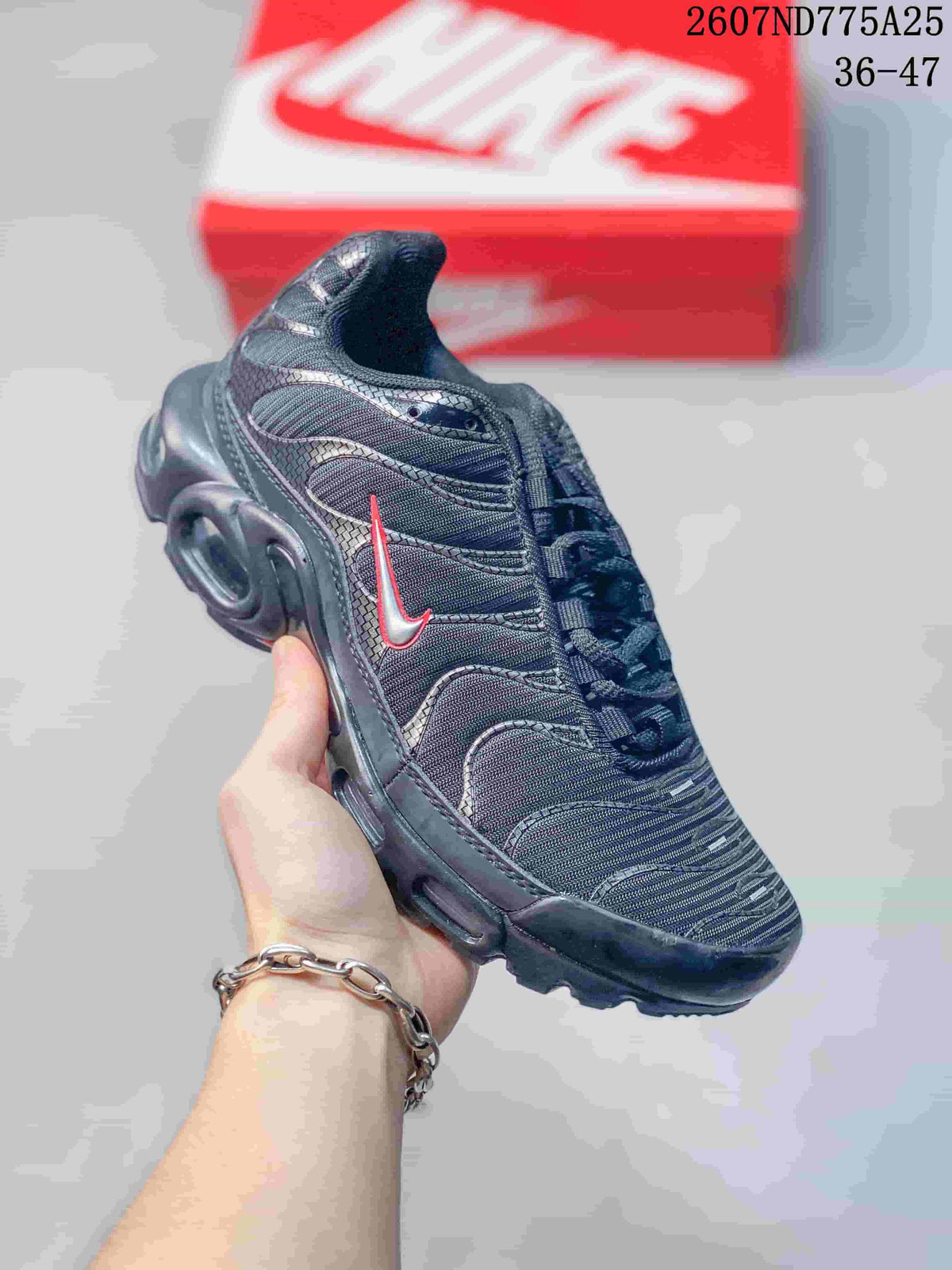 Angled side view of a black Nike Air Max Plus TN sneaker showing the red-accented Swoosh, layered upper, and sculpted midsole; overlay text shows code 2607ND775A25 and size range 36–47.