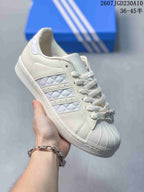 Angled side view of the white Adidas Superstar sneaker showing the shell toe, quilted stripe panel, and Superstar side text.