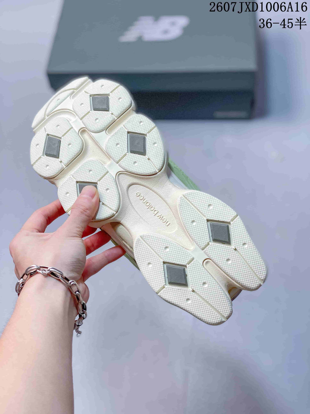 Bottom view of the cream-colored outsole showing segmented tread sections and dark square inserts; a New Balance box is in the background.