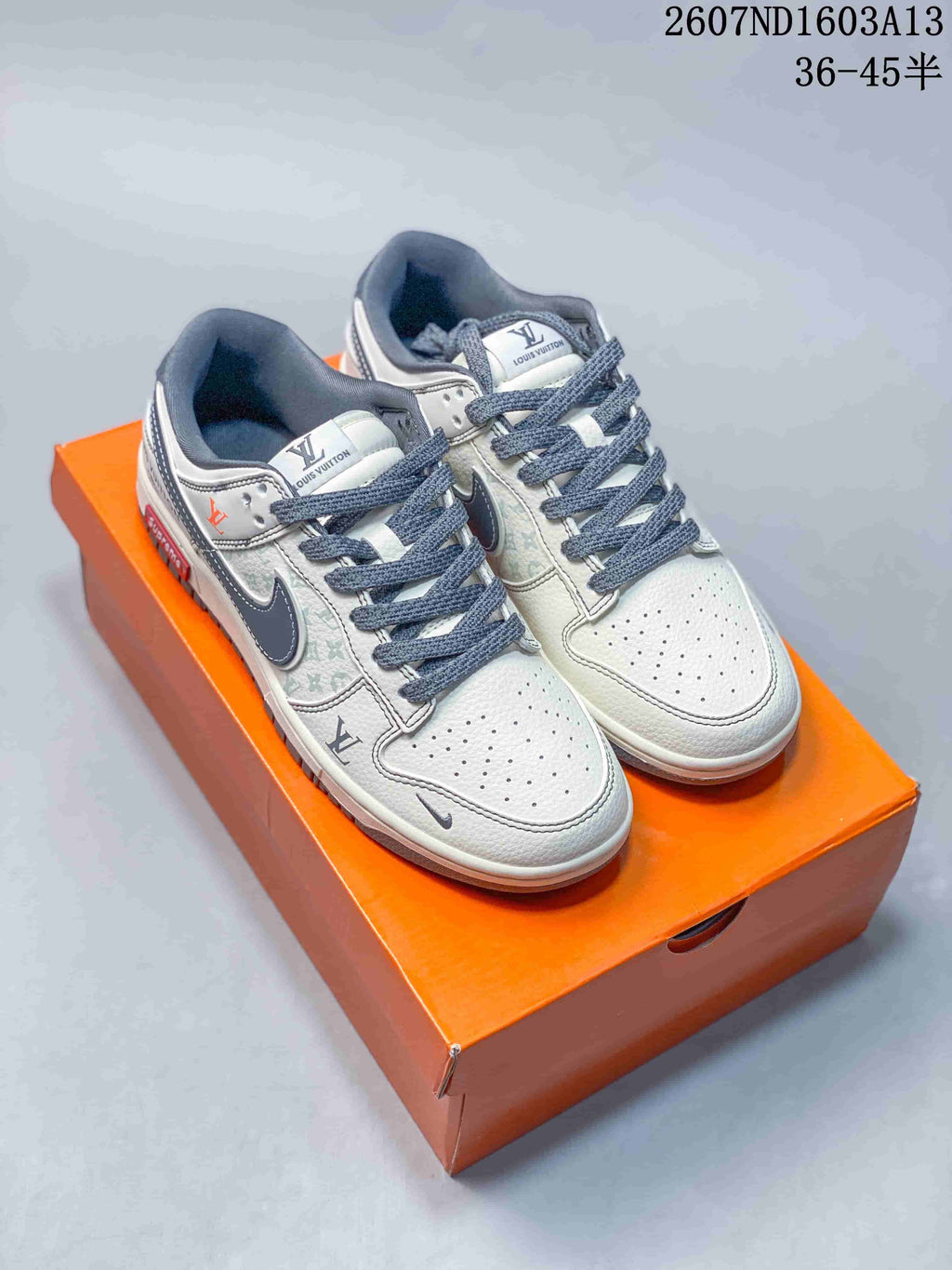 Pair of white and grey Nike SB Dunk Low sneakers displayed on an orange box, with dark grey laces and monogram-style panel detailing.