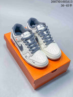 Pair of white and grey Nike SB Dunk Low sneakers displayed on an orange box, with dark grey laces and monogram-style panel detailing.