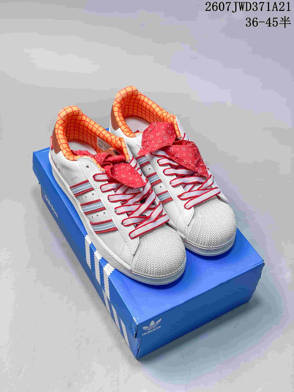 Pair of white Adidas Superstar Supermodified sneakers with red patterned laces, red and light blue side stripes, and a blue shoe box.