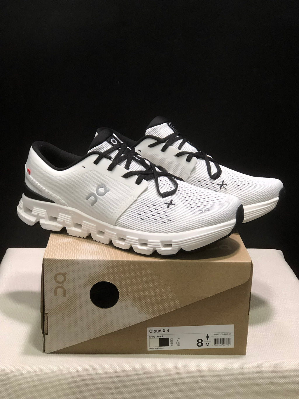 Pair of Ivory and Black On Cloud X4 sneakers displayed on their shoe box.