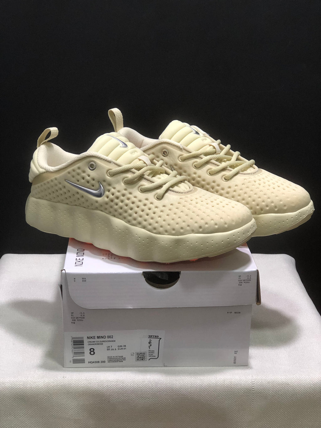 Pair of cream Nike Mind 002 sneakers displayed on a labeled shoebox, showing the textured upper and silver side branding.