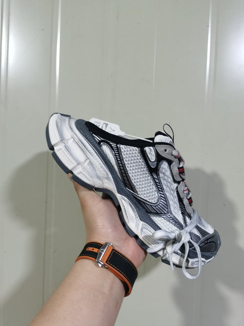 Close-up side view of a grey, white and black 3XL sneaker with layered panels, textured mesh and red lace-area detailing.