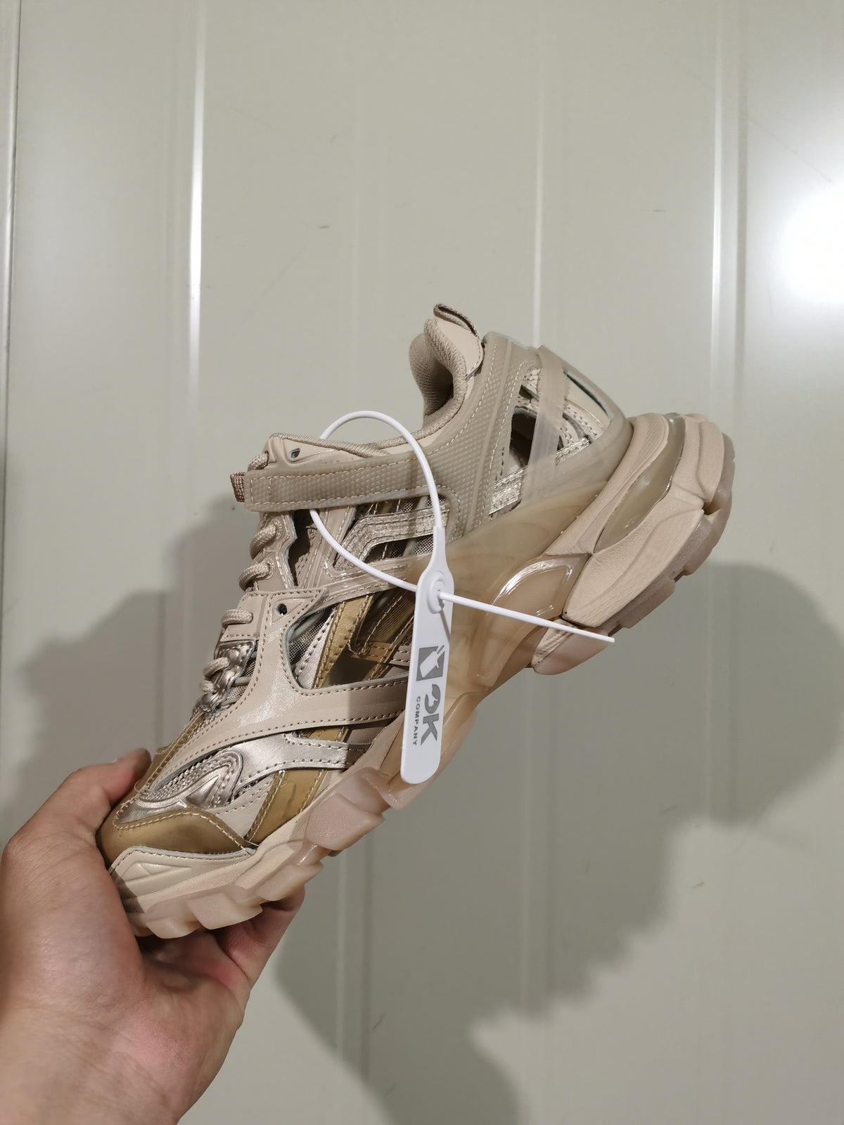 Light apricot sneaker held in hand, showing layered side panels, open sections, and a chunky outsole.