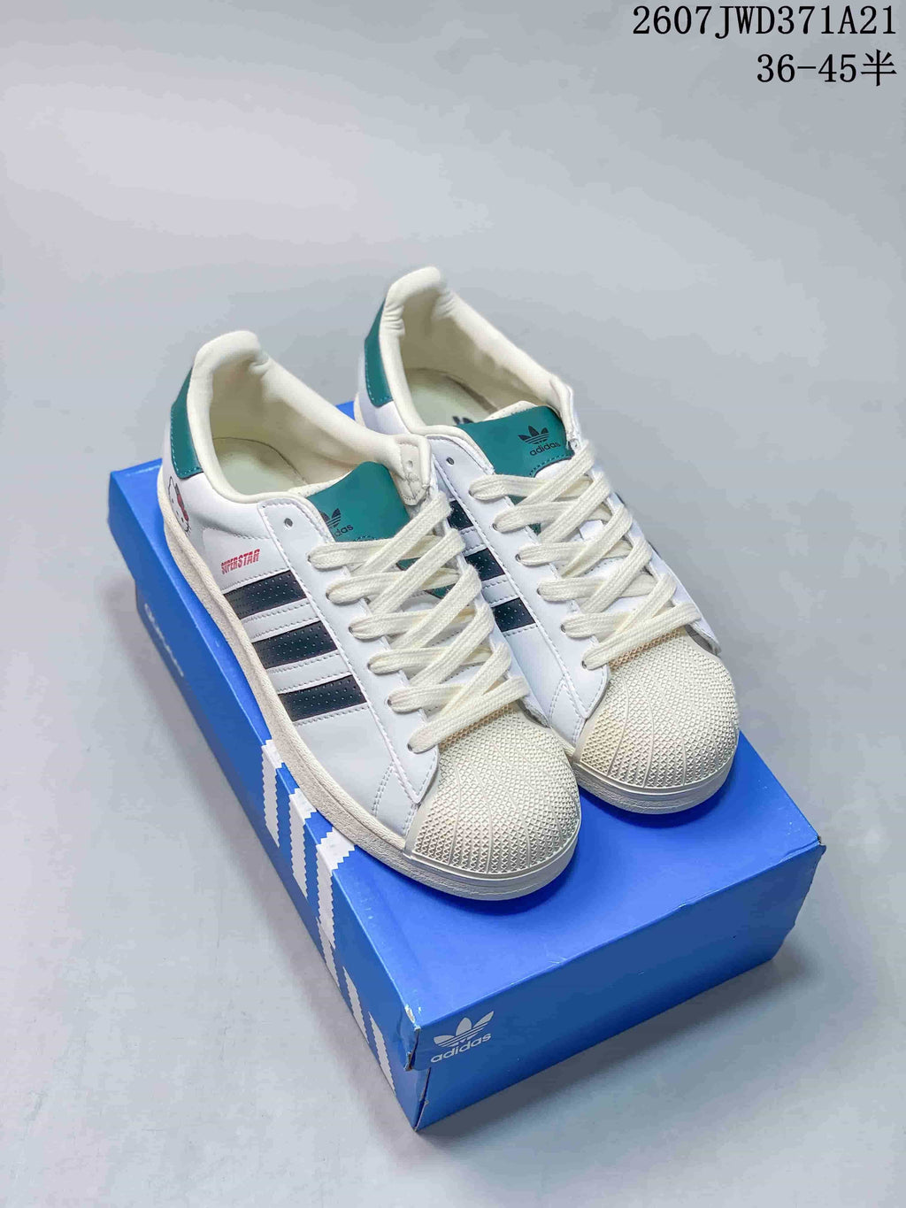 Pair of white Adidas Superstar sneakers with dark blue striped accents, teal tongue and heel details, and textured shell toes on a blue Adidas shoebox; overlay states 2607TWD371A21 and sizes 36–45 with half sizes.