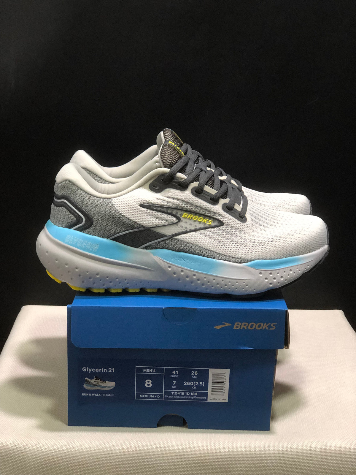 Side view of the Brooks Glycerin 21 pair above its box, showing the white and grey upper, black laces, blue heel accent, and yellow outsole details.