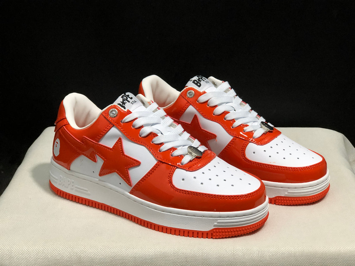 Three-quarter view of the white and orange BAPE Sta sneaker pair with visible side stars and perforated toe boxes.