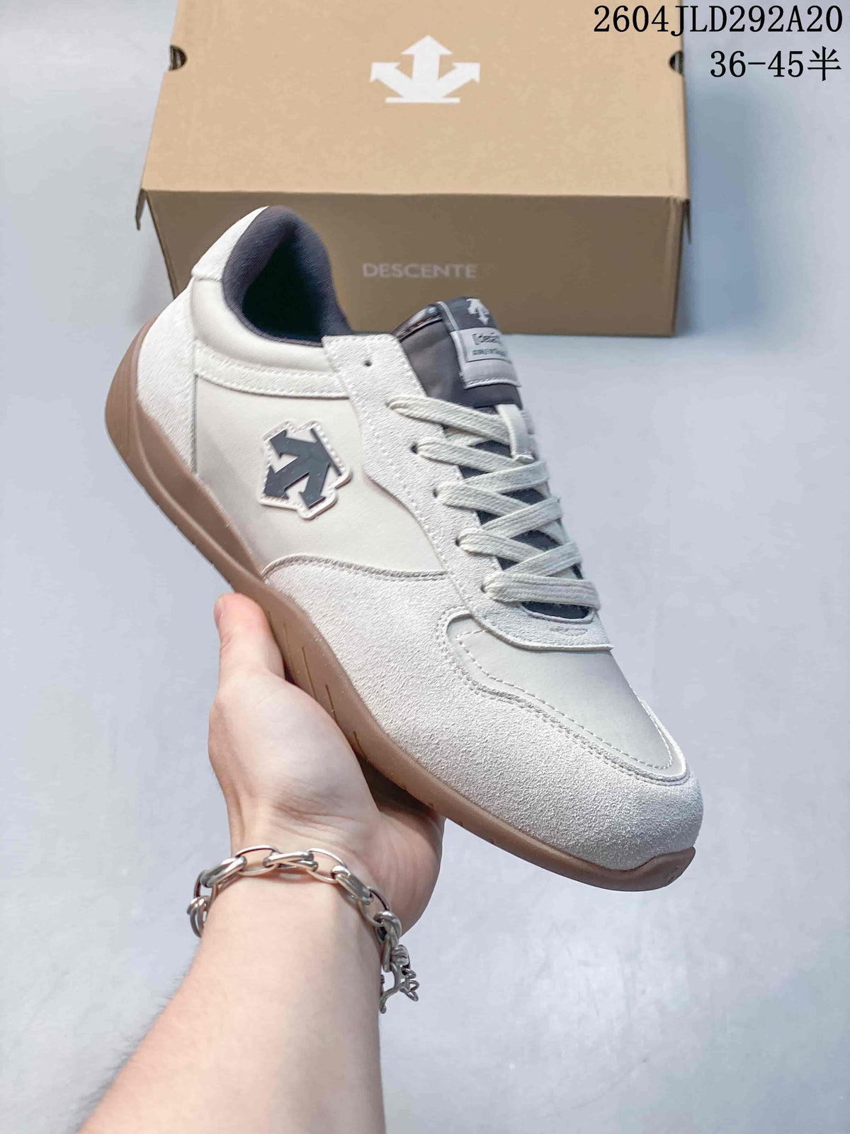 Angled side view of a cream Descente Chron Tempo Slim sneaker with a dark side logo and gum-colored sole.