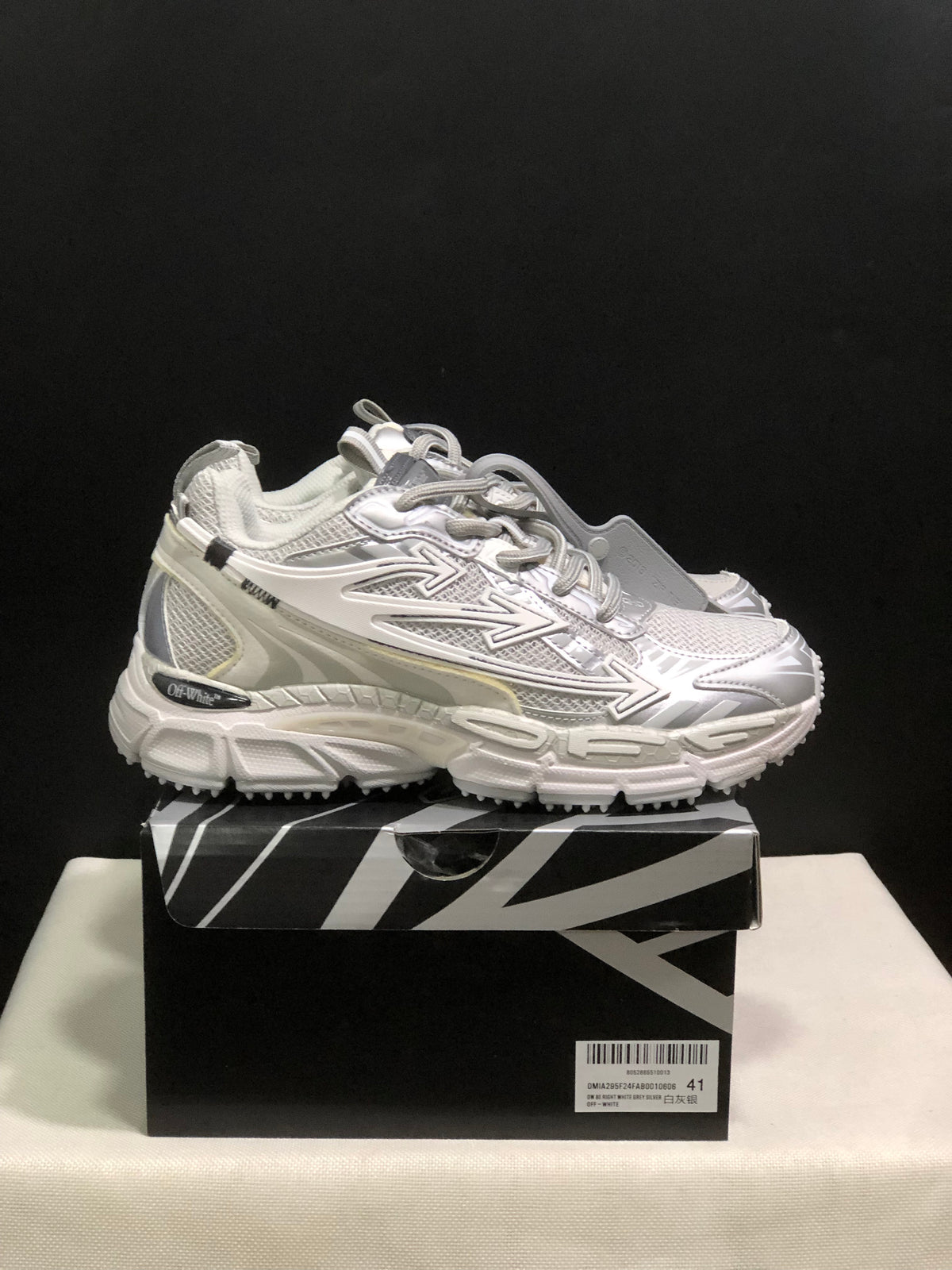 Side view of the white and grey sneakers on the box, showing layered panels, directional side graphics, and chunky soles.
