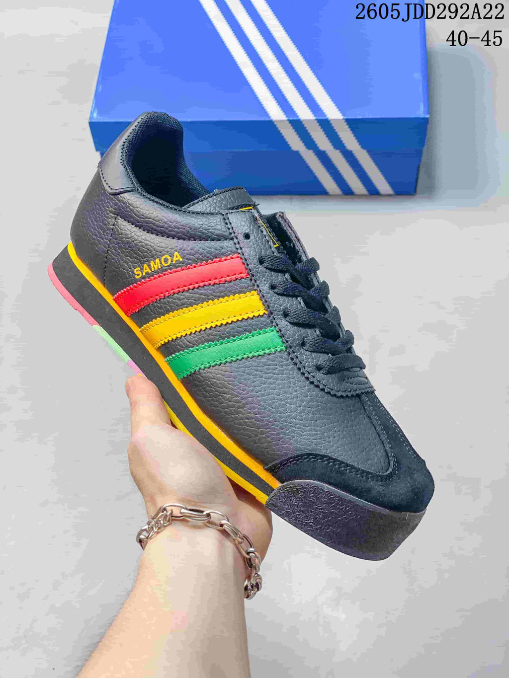 Dark Adidas Samoa sneaker with yellow, red and green side stripes, photographed in front of a blue shoebox.