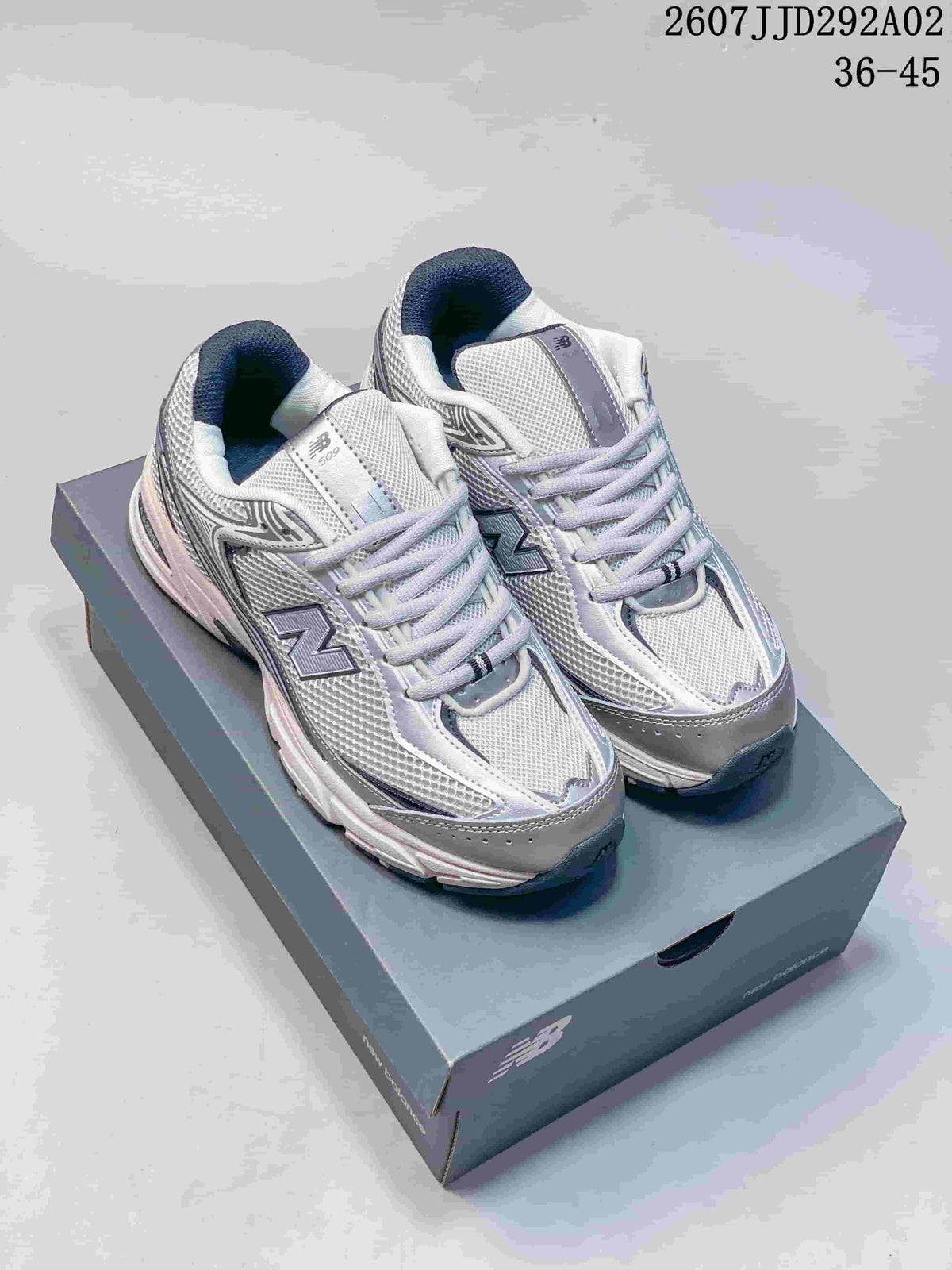 Pair of white and grey New Balance 509 sneakers displayed on a shoebox, with style code JJD292A02 and size range 36-45 visible.