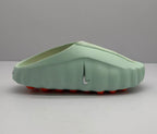 Side profile of a light green Nike slide showing the perforated upper, silver Swoosh-style emblem, and orange outsole elements.
