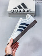 Side profile of a white Adidas sneaker with black three-stripe detailing, gray toe panel, white laces, and a brown outsole.