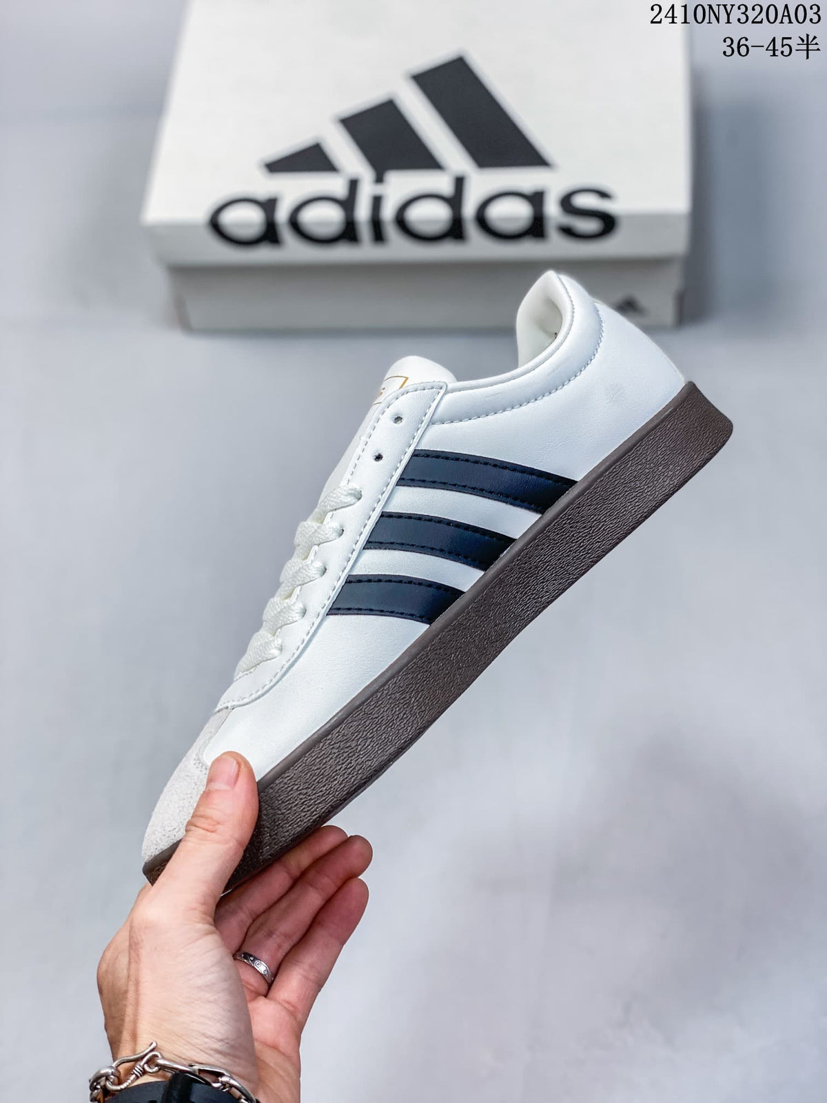 Side profile of a white Adidas sneaker with black three-stripe detailing, gray toe panel, white laces, and a brown outsole.