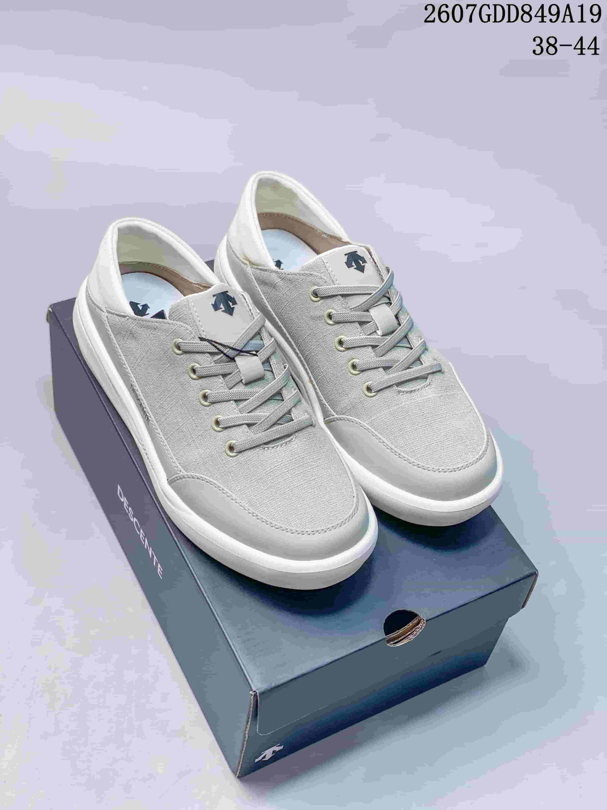 Pair of white and light gray Descente low-top sneakers displayed on a branded shoebox; style code 2607GDD849A19 and size range 38–44 are visible.