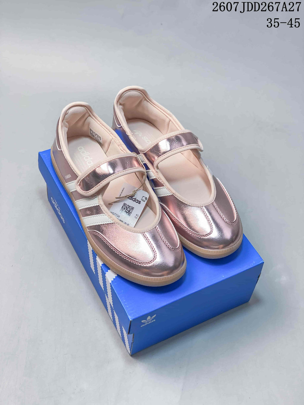 Pair of metallic pink adidas Samba Jane sneakers displayed on a blue shoebox, showing the Mary Jane straps and gum-colored soles.