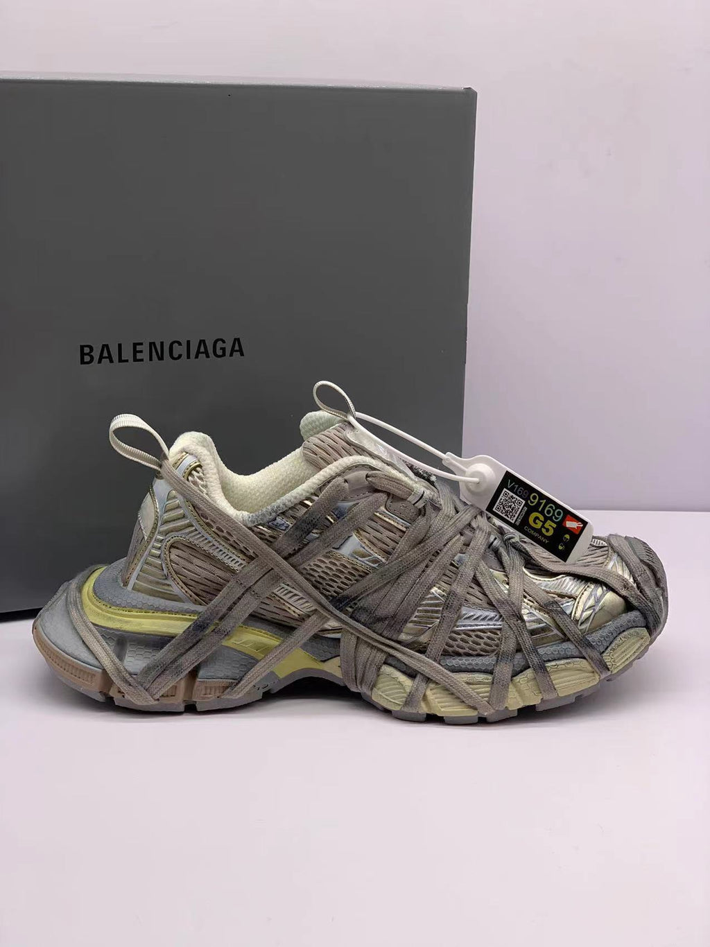 Side view of champagne-toned Balenciaga 3XL-style sneakers with layered panels, overlapping laces, and a branded box in the background.