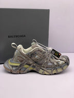 Side view of champagne-toned Balenciaga 3XL-style sneakers with layered panels, overlapping laces, and a branded box in the background.