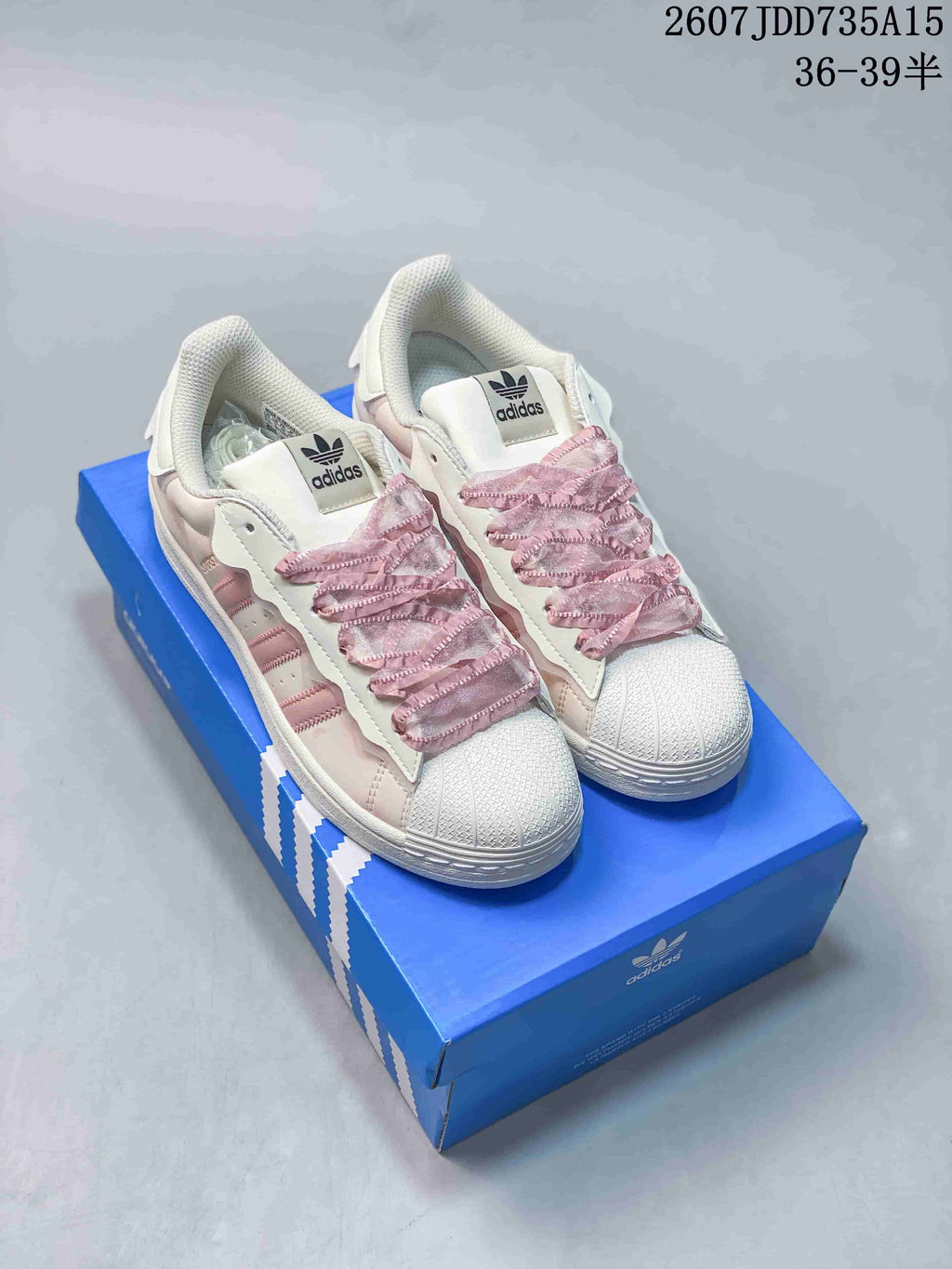 Pair of white and pink adidas Superstar sneakers displayed on a blue shoebox, with the shell toes and ruffled pink laces visible.