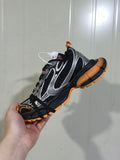 Side view of black sneakers with silver layered detailing and orange outsole accents.