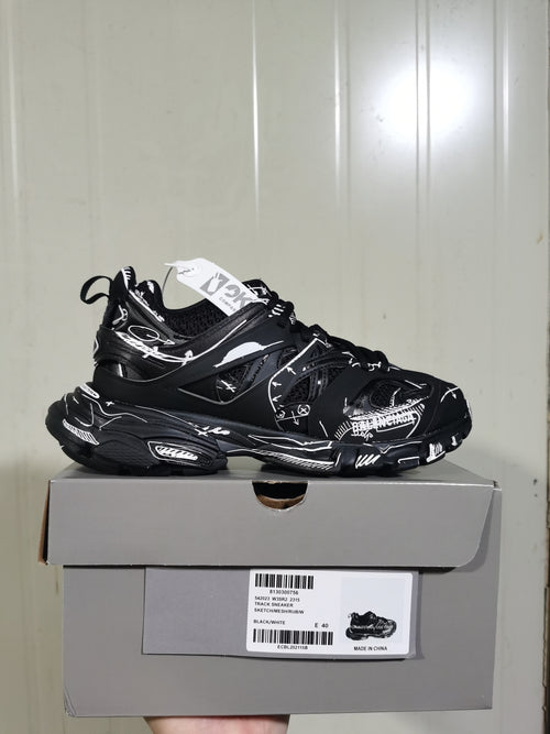 Black and white track sneaker displayed on its box, showing the layered upper, graphic detailing, chunky sole, and box label.