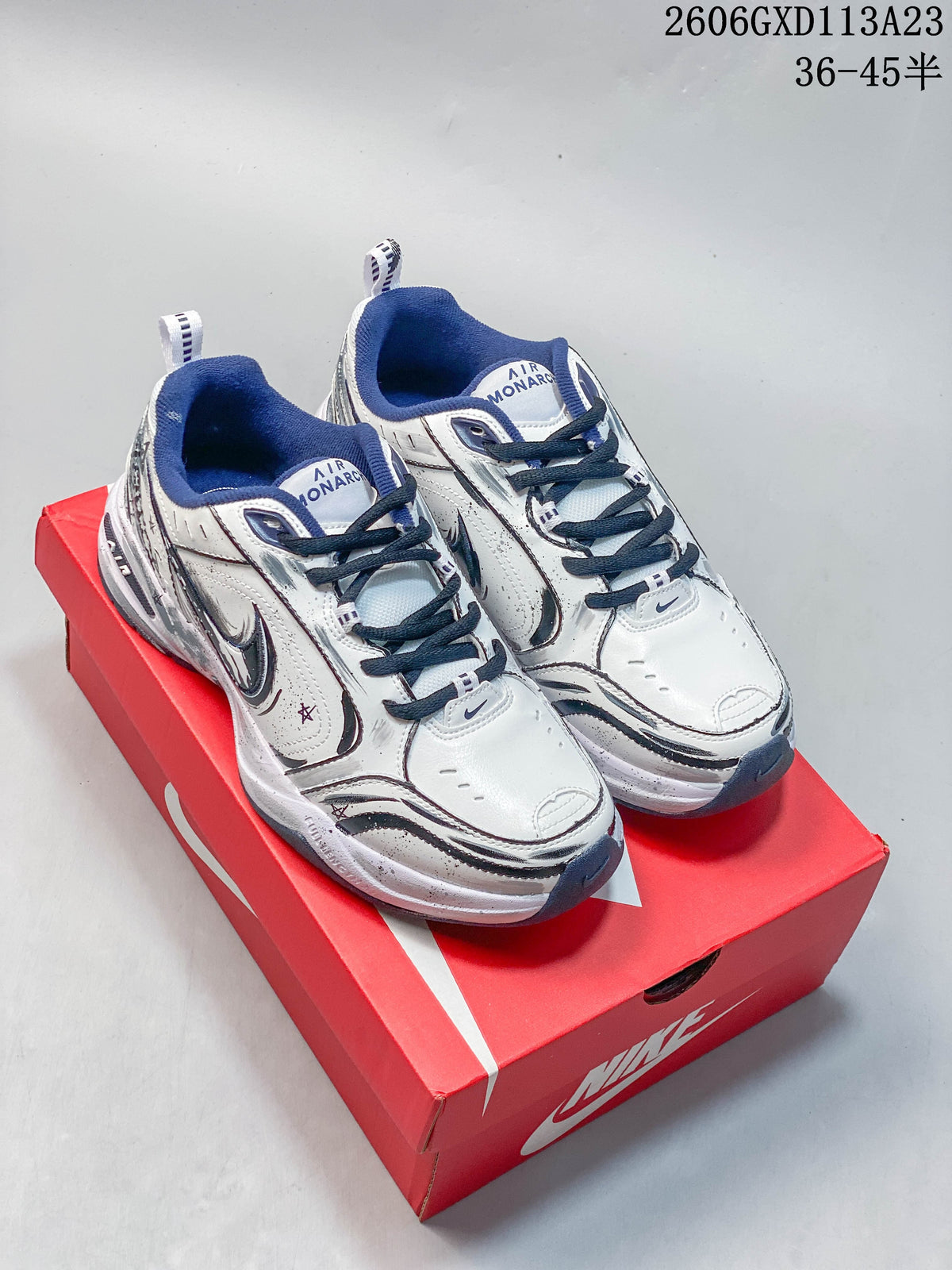 Pair of white Nike Air Monarch IV sneakers with navy and silver details, shown on a red shoebox.