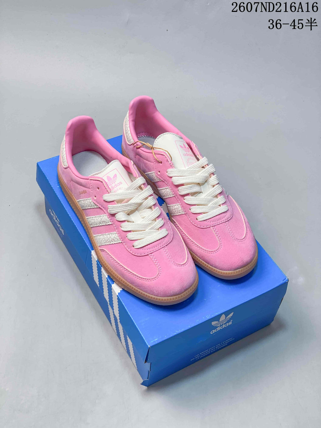 Pair of pink and white adidas Samba sneakers displayed on a blue shoebox, with a gum-colored outsole visible.