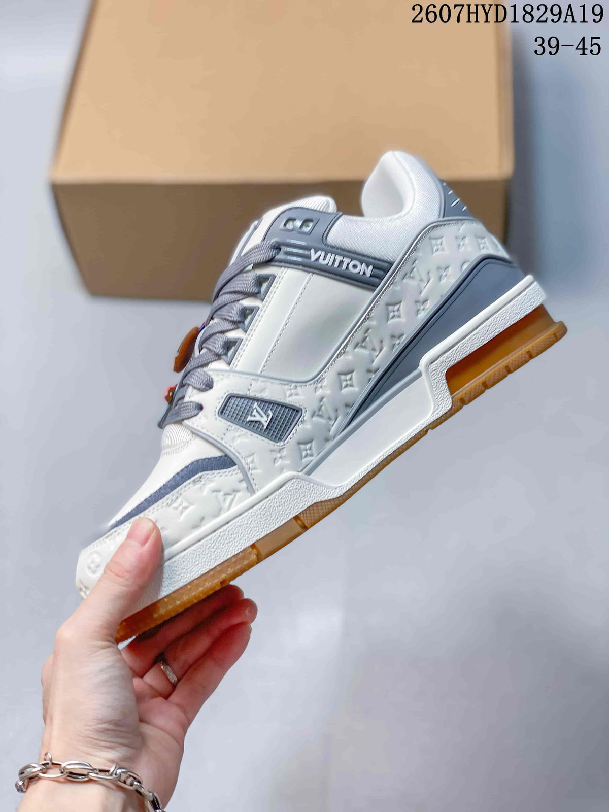 Side view of a white and gray low-top sneaker showing layered panels, embossed motif detailing, gray laces, and a gum-colored outsole.