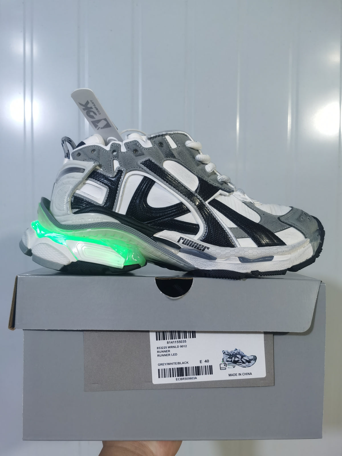 Puma 9012 sneaker displayed on its box, showing a green illuminated heel accent and the grey, white, and black colorway.