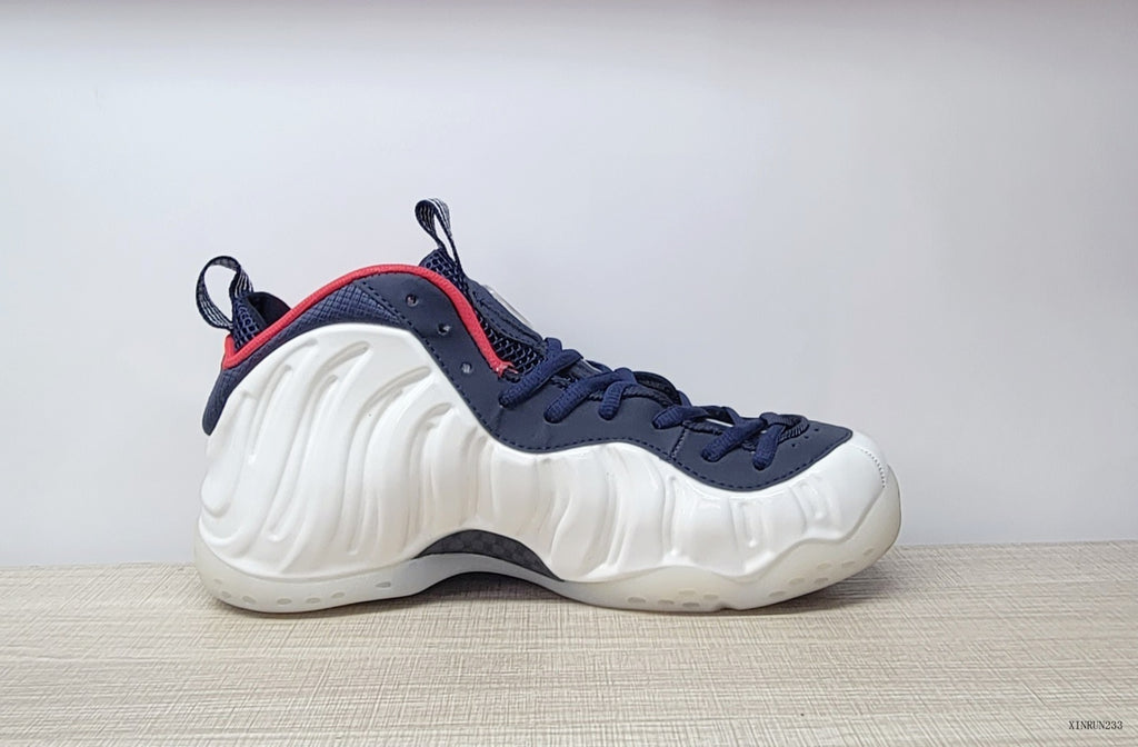 Side profile of a white Nike Air Foamposite One sneaker with navy laces and red collar trim.